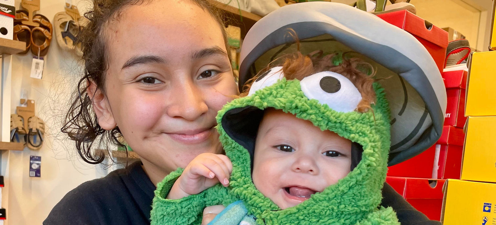 Person holding a baby dressed in a green Oscar the Grouch costume with a bib in a store setting.